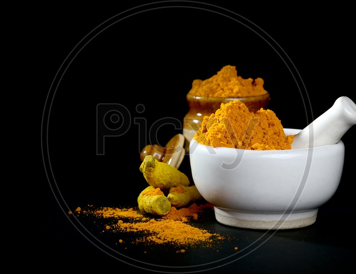 Turmeric powder in mortar with pestle and roots with clay pot on black background