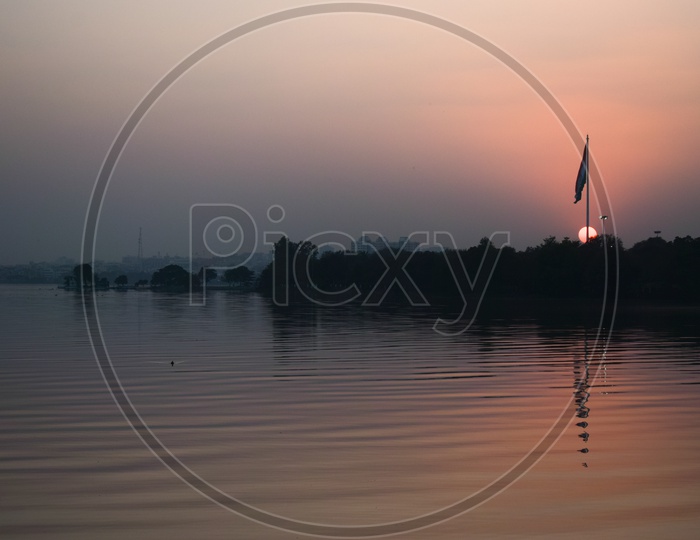 Silhouette Of Flag pole And Its Reflection In Water Surface Over a Sunset Sun Background