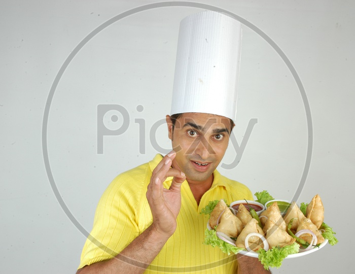 An Indian Chef  In Kitchen Apron And Cap Holding Samosas Plate With an Expression on an Isolated White Background