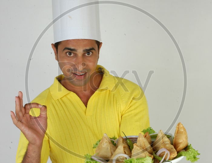 An Indian   Chef   In Kitchen Apron And Cap Holding Samosas Plate With an Expression on an Isolated White Background