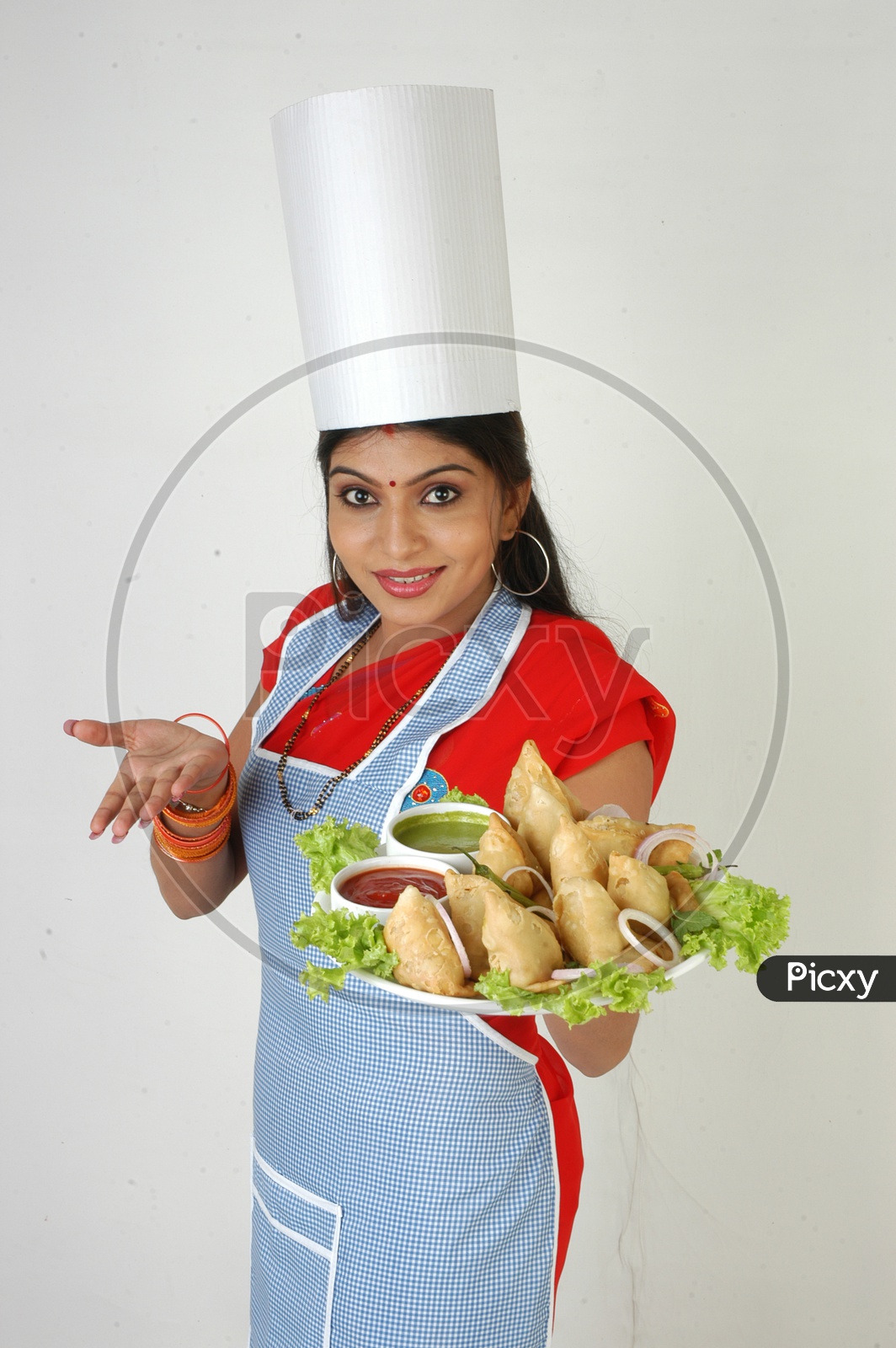 Image Of An Indian Woman Chef In Kitchen Apron And Cap Holding Samosas Plate With An Expression 