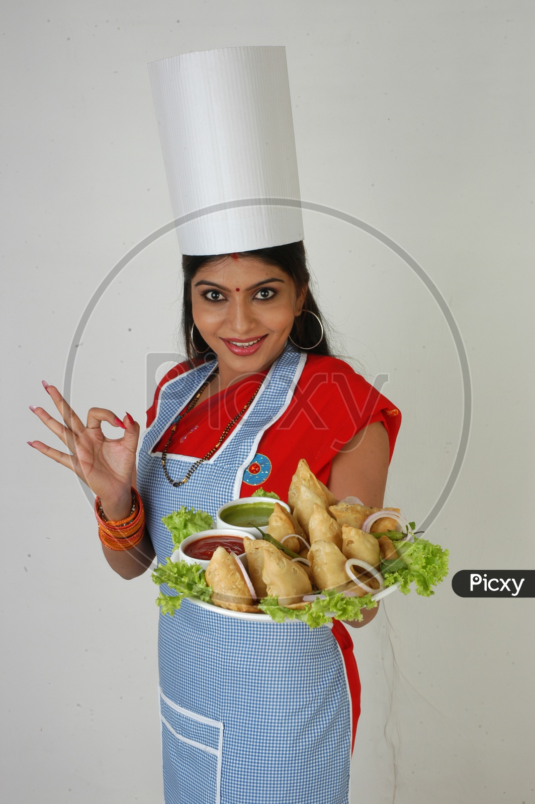 Image of An Indian Woman Chef In Kitchen Apron And Cap Holding Samosas Plate With an Expression