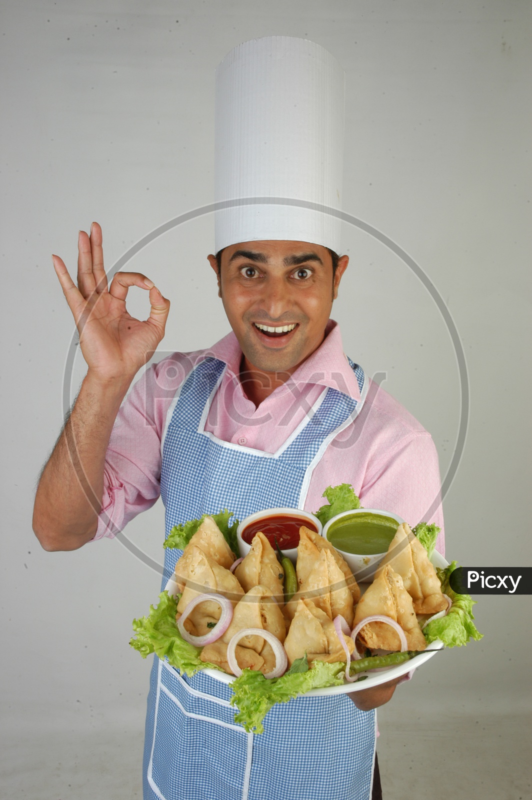 Image Of An Indian Chef In Kitchen Apron And Cap Holding Samosas Plate With An Expression On An 