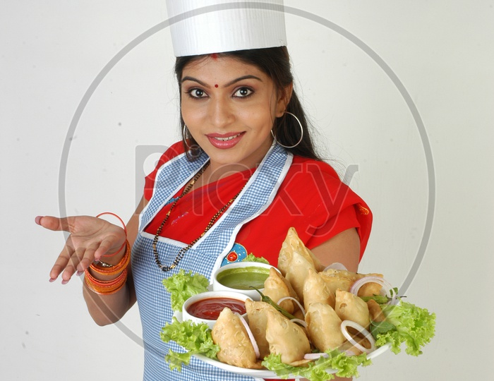 An Indian Woman    Chef   In Kitchen Apron And Cap Holding Samosas Plate With an Expression on an Isolated White Background