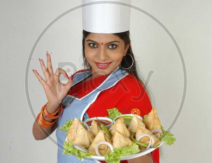 Image of An Indian Woman Chef In Kitchen Apron And Cap Holding Samosas Plate With an Expression