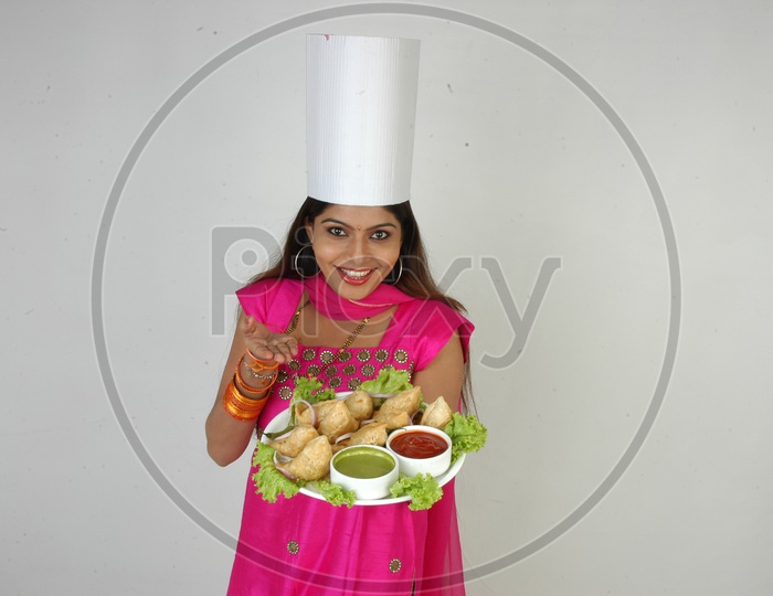 An Indian Woman  Chef  In Kitchen Apron And Cap Holding Samosas Plate With an Expression on an Isolated White Background