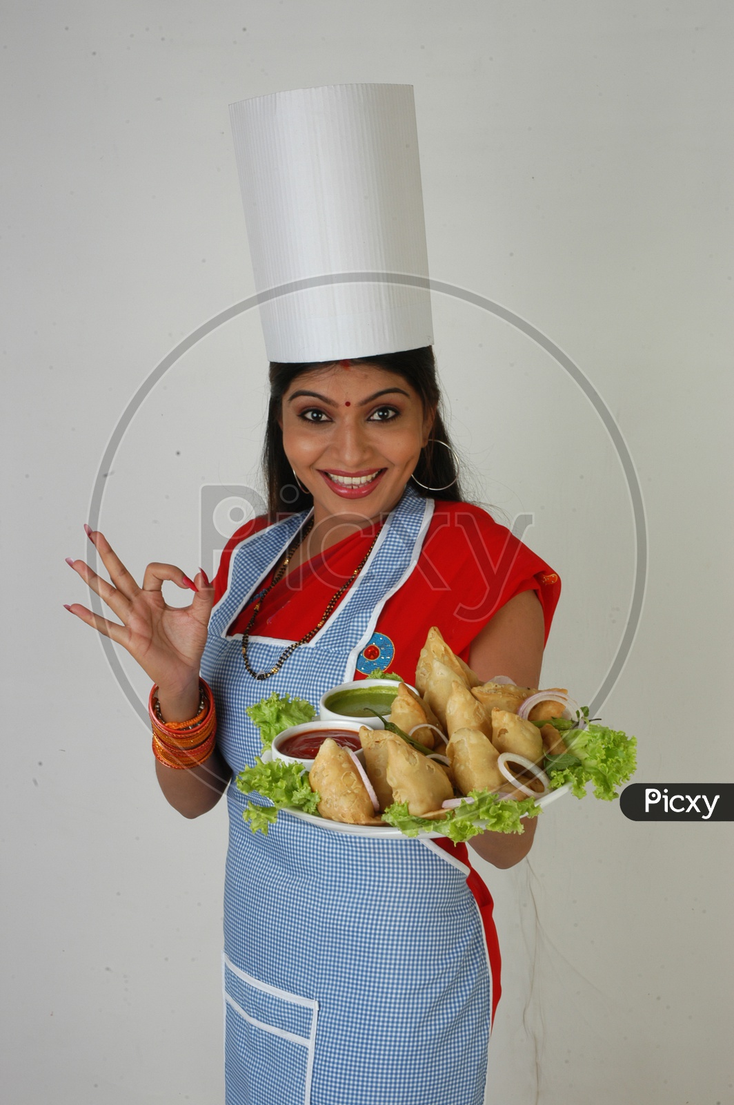 Image of An Indian Woman Chef In Kitchen Apron And Cap Holding Samosas Plate With an Expression