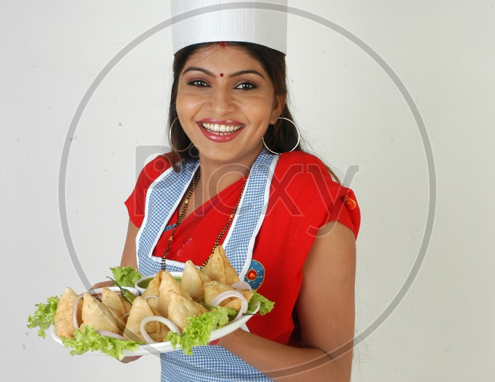 An Indian Woman    Chef   In Kitchen Apron And Cap Holding Samosas Plate With an Expression on an Isolated White Background