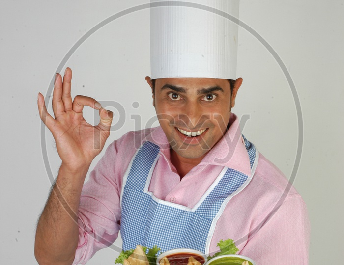 Image Of An Indian Chef In Kitchen Apron And Cap Holding Samosas Plate With An Expression On An 
