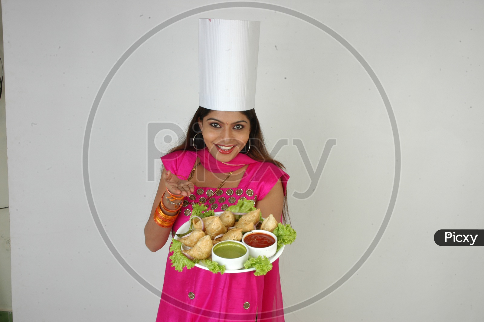 An Indian Woman  Chef  In Kitchen Apron And Cap Holding Samosas Plate With an Expression on an Isolated White Background