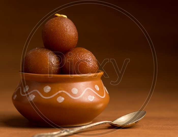 Indian Sweet or Dessert or Savoury Gulab Jamun  Served in an Elegant  Clay  Pot with a Spoon Besides Pot   On an Isolated Wooden Background