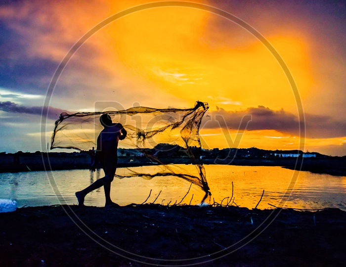 Silhouette of Fisher Man Throwing Fishing Net in a Fishing Pond with Sunset Sky In Background