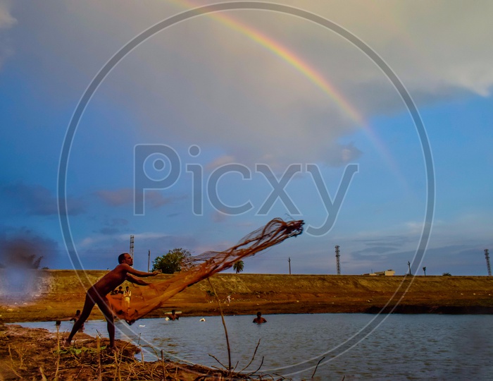 A Fisher Man Throwing Fishing Net into a Fishing Pond  With  Rainbow In Background