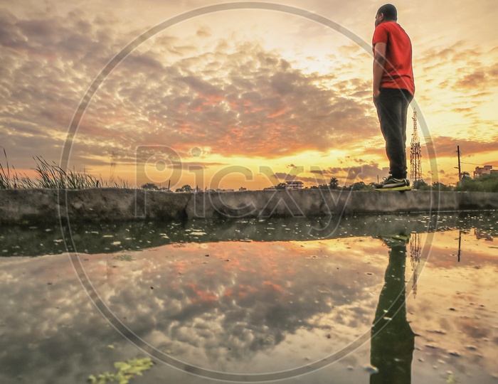 A Man Enjoying Nature or Alone Looking to Space  And his Reflection On Water  With Sunset Sky In Background