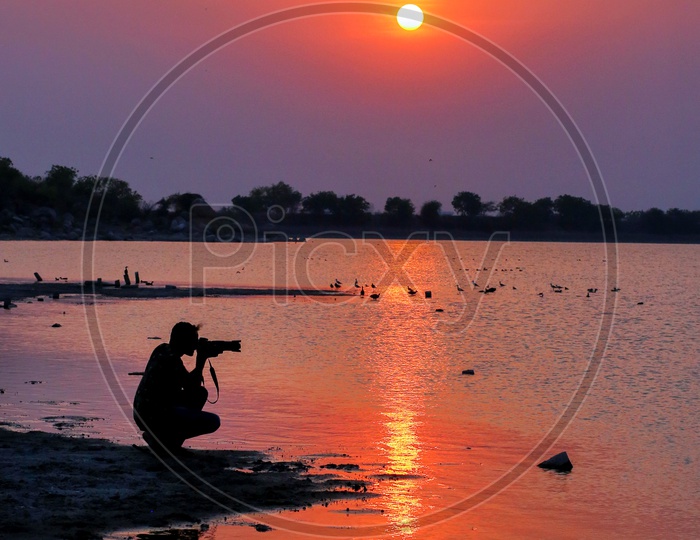 Silhouette Of a Photographer Shooting At Bank Of a Lake With Sunset Sky In Background