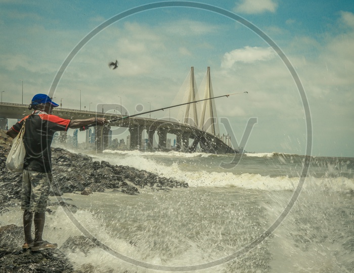 A Man Fishing On The Rock Beach in Arabian Sea  With Bandra Worli Sea Link In Background