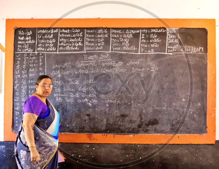 A Govt teacher standing near a blackboard.