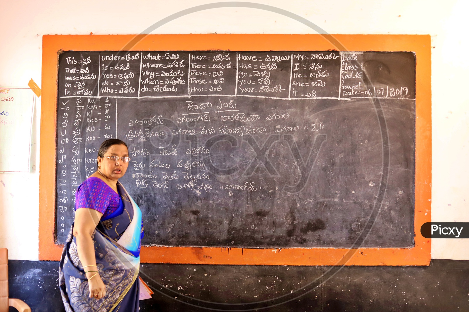 A Govt teacher standing near a blackboard.