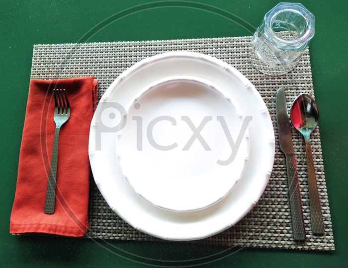 Dinner plate, quarter plate, spoon, knife, fork & water glass set up on table mat.