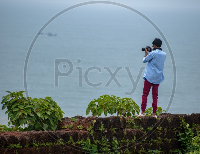 Young man/boy standing on the wall and clicking pictures from Chapora Fort.