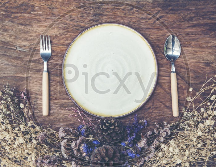 Empty ceramic plate with spoon and fork, dry flowers on wooden background