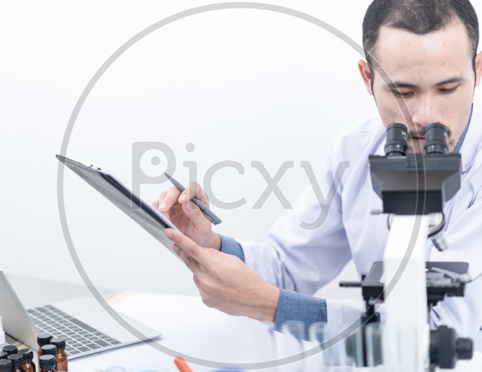 Asian Male Scientist Updating the Sample Test Results from Microscope in Tablet and Laptop