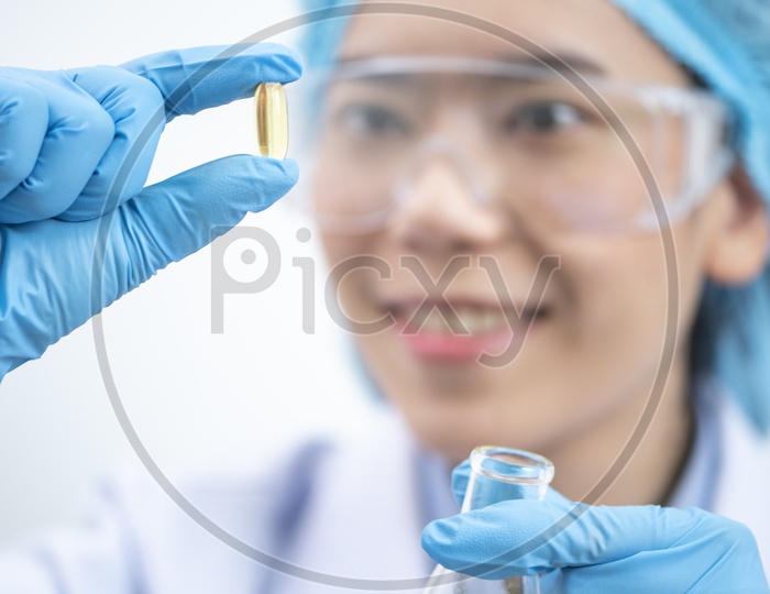 Young attractive female scientist with protective eyeglasses and mask holding a yellow transparent pill with fingers in gloves in the pharmaceutical research laboratory