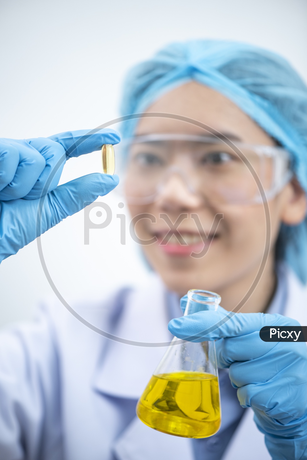 Young attractive female scientist with protective eyeglasses and mask holding a yellow transparent pill with fingers in gloves in the pharmaceutical research laboratory