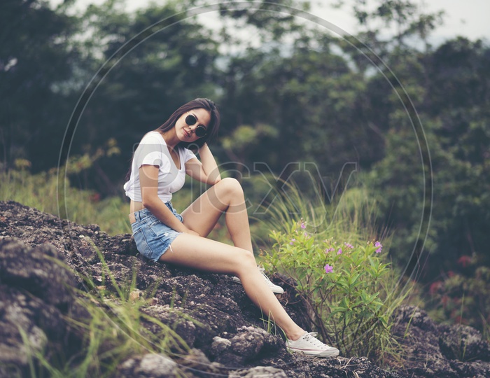 Asian Girl In Modern Wear Posing by Sitting on a rock in outdoor Background