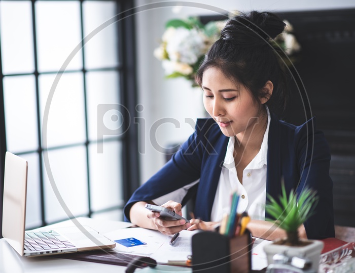 Young Asian Businesswoman using Mobile or Smartphone while working on Laptop at Workplace