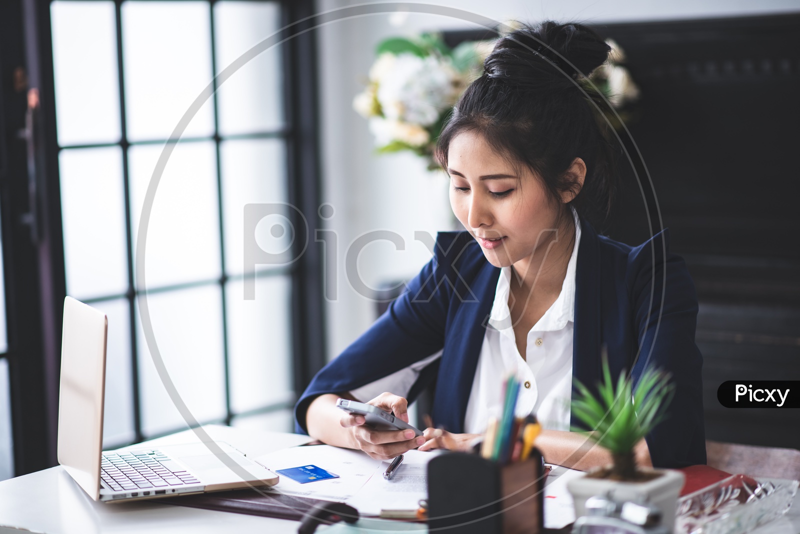 Young Asian Businesswoman using Mobile or Smartphone while working on Laptop at Workplace