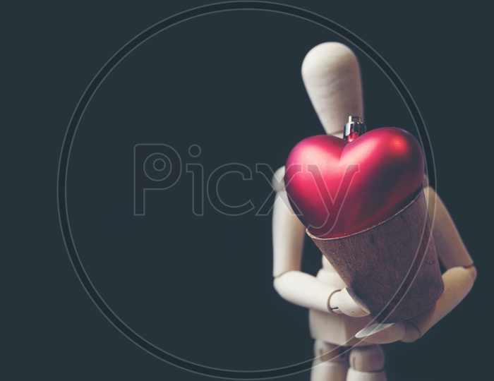 Close up of a wooden human shaped toy holding a soft fur heart shaped toy in wooden cup with black background