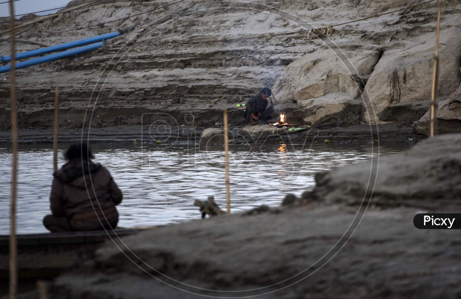 A Fisherman Fishing In The Brahmaputra River And A Man Warm Himself In Bonfire, In Guwahati,