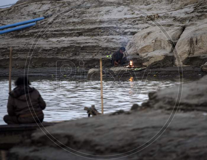 A Fisherman Fishing In The Brahmaputra River And A Man Warm Himself In Bonfire, In Guwahati