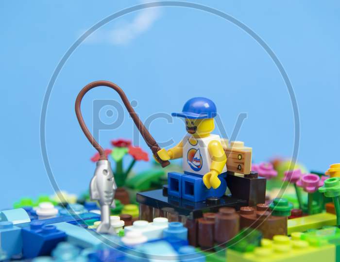 Florianopolis, Brazil. September 20, 2020: Happy Man Minifigure In Tank Top And Cap Sitting On A Wooden Bench Fishing For A Fish. Fishing Is A Sport Practiced Worldwide And By People Of All Ages.