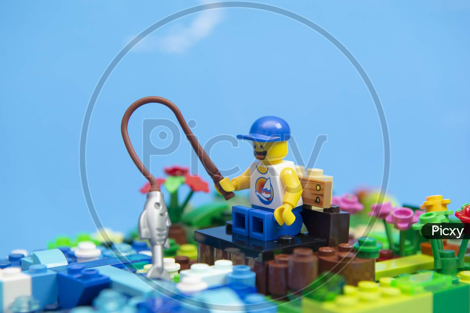 Florianopolis, Brazil. September 20, 2020: Happy Man Minifigure In Tank Top And Cap Sitting On A Wooden Bench Fishing For A Fish. Fishing Is A Sport Practiced Worldwide And By People Of All Ages.