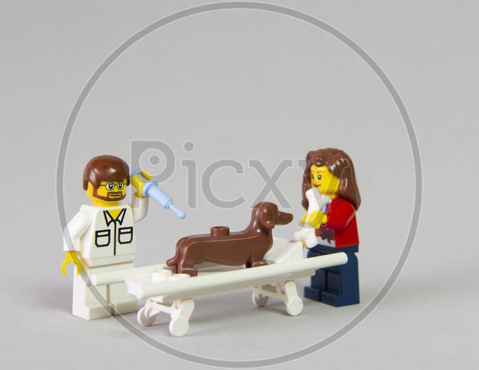 Florianopolis, Brazil. September 19, 2020: Vet Minifigure Giving Injection To A Dog While Its Owner Distracts Him With A Bone On White Background. Concept Of Care For Pets. Selective Focus. Dachshund.
