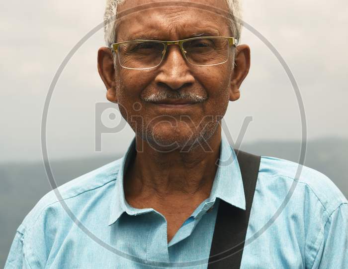 An Old Man, Wearing Spectacles And Blue Shirt, Standing Alone In Nature A Blurry Mountain Background. The Elder Person With A Side Bag On His Shoulder, Looking Towards The Camera. Copy Space For Text.