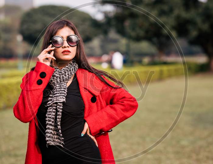 Outdoor Fashion Portrait Of Young Beautiful Fashionable Female Model Wearing Trendy Long Red Winter Coat, Black Suede Ankle Boots, Scarf And Sunglasses.