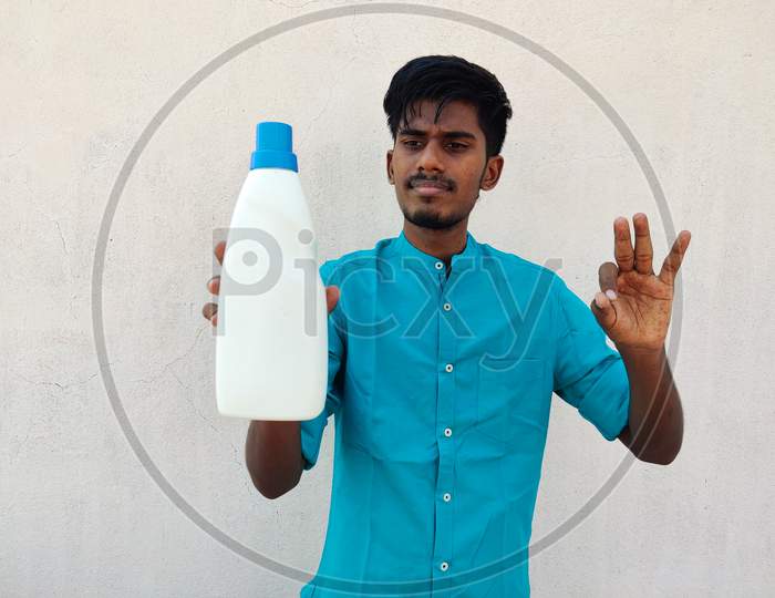 South Indian Man In Cyan Color Shirt Holding White Color Detergent Bottle And Showing Ok Sign.Isolated On White Background.
