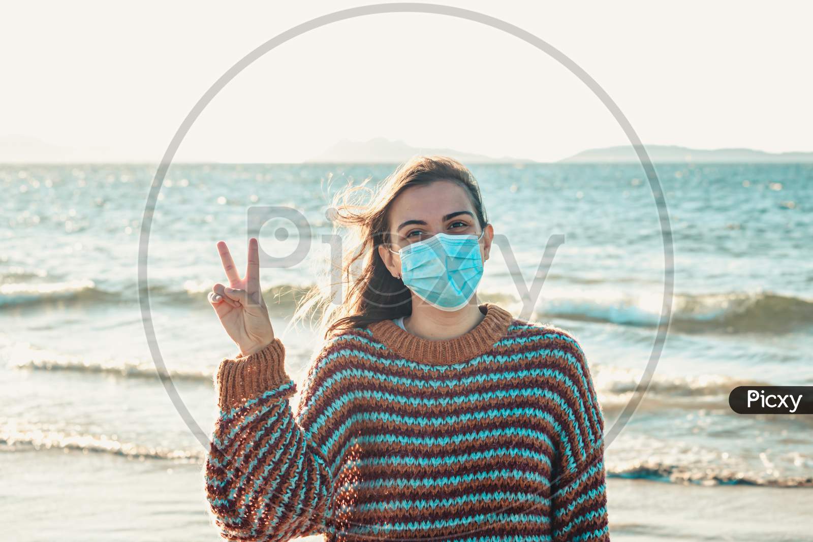 Young Woman On A Sweater On The Beach Using A Surgical Mask And Making The V Symbol During A Bright Day With An Island As The Background