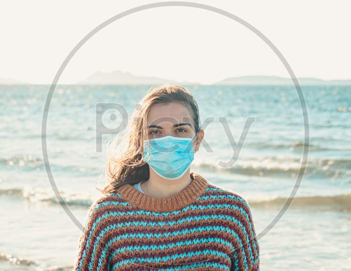 Young Woman On A Sweater On The Beach Using A Surgical Mask During A Bright Day With An Island As The Background