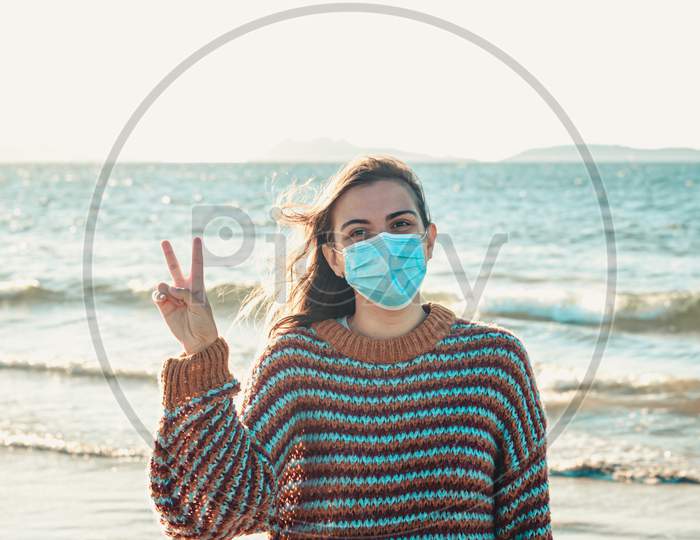 Young Woman On A Sweater On The Beach Using A Surgical Mask And Making The V Symbol During A Bright Day With An Island As The Background