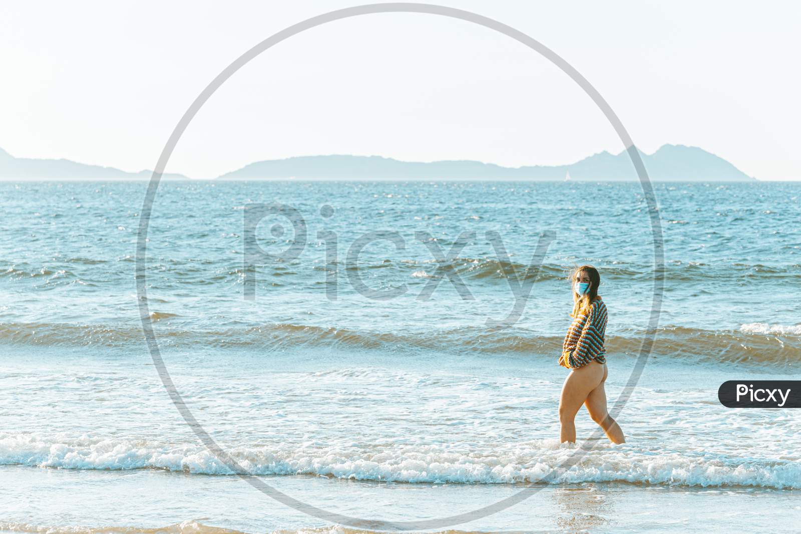 Young Woman Walking On The Sea In The Beach With A Surgical Mask During A Bright Day With A Sweater Put On And Copy Space