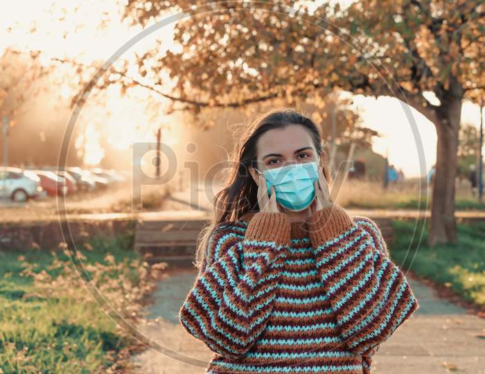Young Woman Wearing A Surgical Mask And An Autumnal Sweater During A Sunset In A Natural Park On Colorful Tones