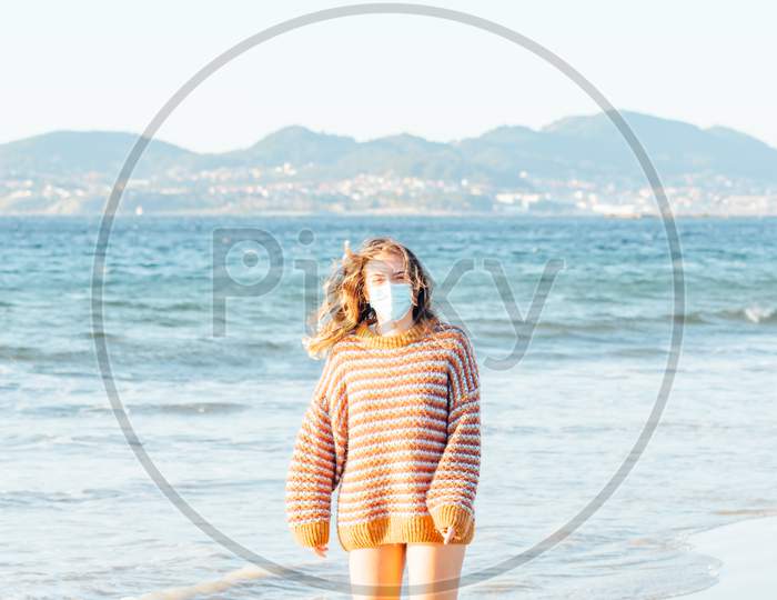 Young Woman On The Beach With A Surgical Mask During A Bright Day Wearing A Sweater With Copy Space And Happy Aptitude