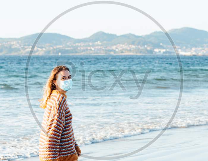 Young Woman Walking On The Sea In The Beach With A Surgical Mask During A Bright Day With A Sweater Put On And Copy Space