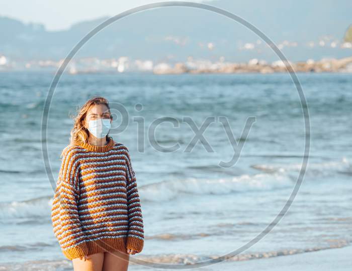 A Young Woman On The Beach With A Surgical Mask During A Bright Day Wearing A Sweater With Copy Space