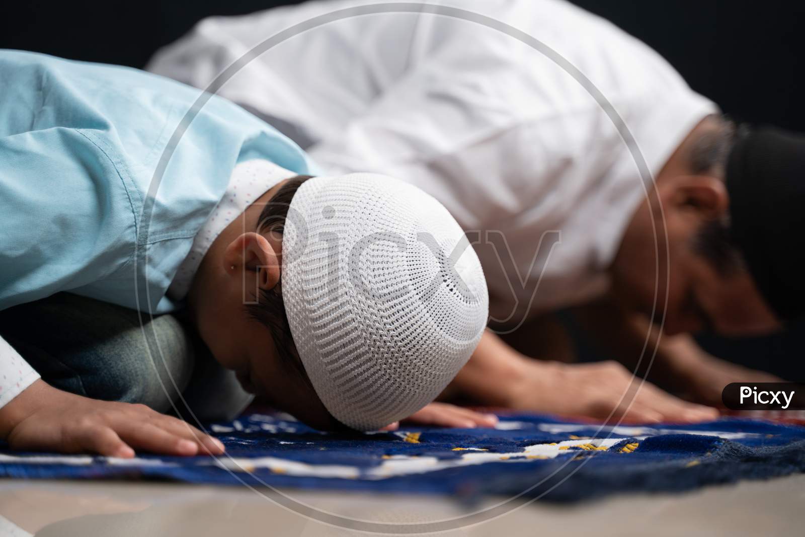 Muslim Father And Son Praying Or Performing Salah While Sitting On Prayer Rug And Touching Head To Mihrab Or Mosque Printed On Rug.
