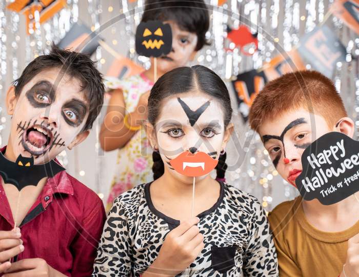 Group Of Kids In Halloween Costumes Making Gesticulating Scary Or Spooky Faces By Holding Booth Sprops On Decorated Background By Looking Into Camera.